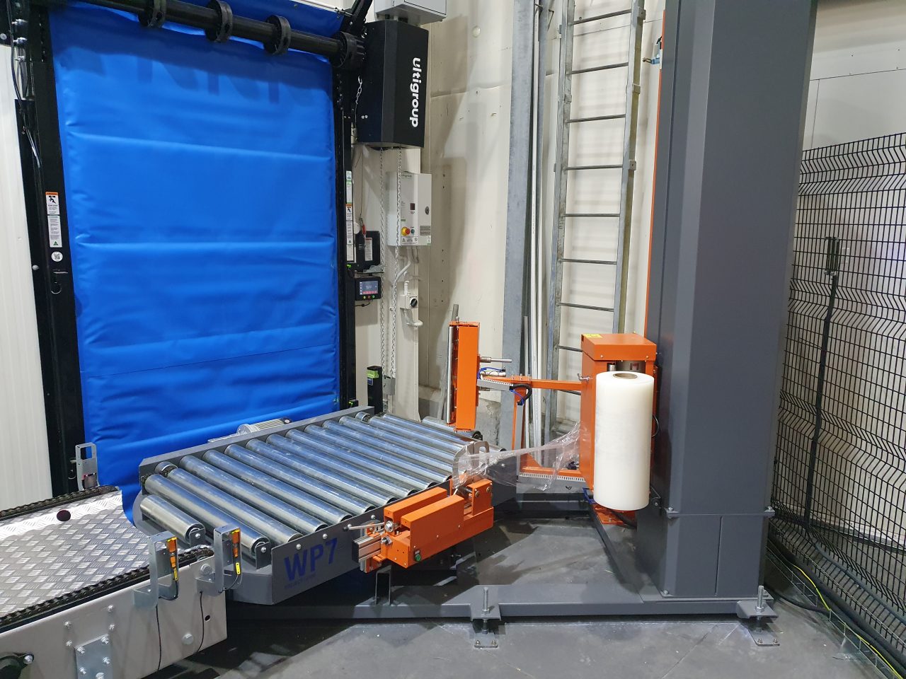 PHS Innovate turntable pallet wrapper on an infeed roller conveyor, with the film carriage clamping the stretch film at the start of a cycle